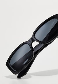Sorte solbriller med en tykk, vinkelret plastikramme og mørke linser. Bemærkelsesværdige træk inkluderer store firkantede former og minimalistisk design.