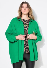 Zelená košile na knoflíky vyrobená z texturované látky, s volným střihem a vyhrnutými rukávy, nošená přes top s leopardím vzorem a legíny.
