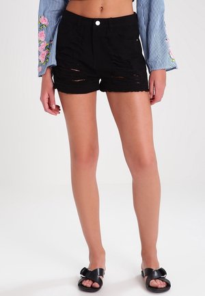 Mujer con pantalones cortos de mezclilla negros desgarrados, una camiseta azul a rayas con bordados florales en las mangas y sandalias negras con hebillas.
