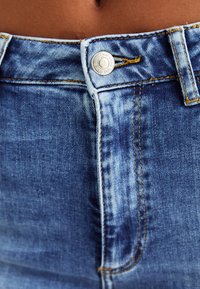 Augstas jostas denim bikses ar gaišu mazgāšanu un izbalējušām vietām. Iekļauj metāla pogu aizdari, redzamu šūšanu un klasisko piecu kabatu dizainu.