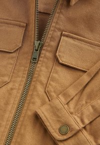 Bruine textieljas met een rits aan de voorkant, twee borstzakken en een knoopsluiting aan de mouwen. De stof heeft een gestructureerde afwerking met een stevige uitstraling.