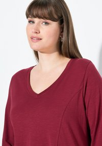 Burgunderfarbener Langarmtop mit V-Ausschnitt, aus sanftem Material und mit dezenten Nahtdetails, vor einem neutralen Hintergrund.