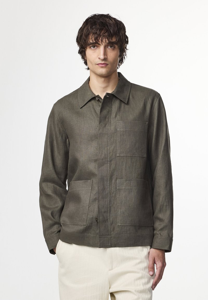 Groene textuurjack met een kraag, voorknopsluiting en twee opgestikte zakken. Draagt over lichtgekleurde broek, tegen een neutrale achtergrond.