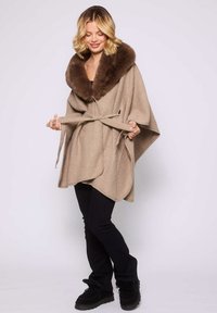 Femme nouant une ceinture sur une cape beige avec un grand col en fourrure marron, portant un pantalon noir et des bottes d'hiver noires, se tenant sur un fond gris clair.