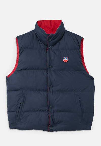 Chaleco acolchado azul marino con acentos rojos, cuello alto, cierre frontal de botones y bolsillos laterales. Presenta un logotipo de la marca en el pecho.