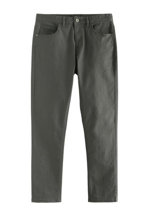 Noormees seisab käed ees taskus, seljas beež kapuutsiga dressipluus, tumedad püksid ja valged tossud, ühevärvilise heleda tausta ees.