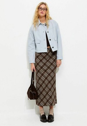 Młoda kobieta o blond włosach i w okularach, ubrana w jasnoniebieską kurtkę, brązową spódnicę w kratę, beżowe skarpetki, czarne buty, trzymająca brązową torbę.