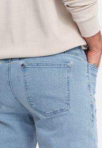 Jasnoniebieskie dżinsy mają dopasowany krój, tylnie kieszenie, subtelne przeszycia oraz postrzępiony dół na jednej nogawce.