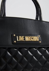 Musta tikattu käsilaukku, jossa sileä nahkainen yläosa, kahdet kahvat ja kultainen "Love Moschino" -logokilpi, joka on kiinnitetty neljällä kultaisella niitillä.