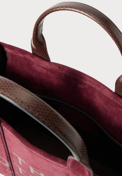 Közelkép egy bordó velúr bevásárlótáskával, barna textúrált bőr fogantyúkkal és látható belső cipzáras zsebbel.