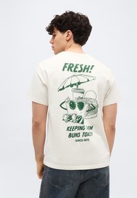 Νεαρός άνδρας που φοράει λευκό T-shirt με πράσινο γραφικό που δείχνει ένα ποτό με γυαλιά ηλίου κάτω από ομπρέλα, ένα μπέργκερ και το κείμενο «ΦΡΕΣΚΟ! ΚΡΑΤΩΝΤΑΣ ΤΑ ΨΩΜΑΚΙΑ ΖΕΣΤΑ.»