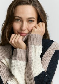 Pull en tricot beige, marine et crème avec un design à rayures. Présente une texture épaisse et des poignets côtelés, partiellement relevé au niveau du col.