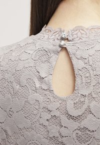 Lys grå blonde-top med rund halsudskæring, blomsterdesign og en nøglehul-udskæring i ryggen med to dekorative, klare knapper.