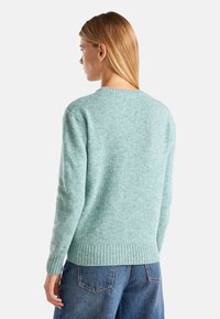 Jasnozielony sweter z dzianiny z okrągłym dekoltem, ściągaczami na mankietach i dole. Miękka tekstura, długie rękawy i luźny krój. Noszony z niebieskimi dżinsami.