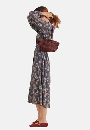 Abito maxi con motivo floreale in blu navy e bianco, vita cintrata, maniche lunghe; abbinato a una borsa intrecciata bordeaux e ballerine rosse.