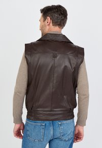 Braune Lederweste mit einem Reverskragen, seitlichen elastischen Einsätzen und einem metallenem Akzent. Getragen über einem leichten Pullover, kombiniert mit blauen Jeans.