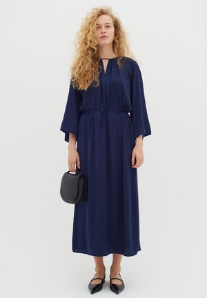 Marineblauwe maxi-jurk van lichte stof met een koord in de taille, keyhole-halslijn en brede mouwen. Gepaard met een zwarte handtas en flats.