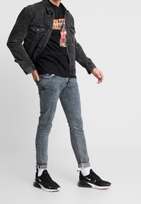 Chaqueta de denim negra sobre una camiseta gráfica negra, jeans ajustados azules y zapatillas deportivas negras con acentos en blanco y puños doblados.