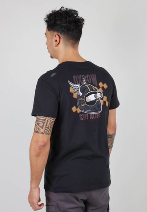 T-shirt en coton noir avec un graphique d'un cochon portant un casque avec des ailes, texte indiquant "OXBOW STAY ALIVE", et des accents colorés en motif à carreaux.