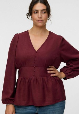 Femme posant la main sur la hanche, portant un chemisier bordeaux à manches longues avec des boutons recouverts de tissu et un jean bleu sur un fond uni.