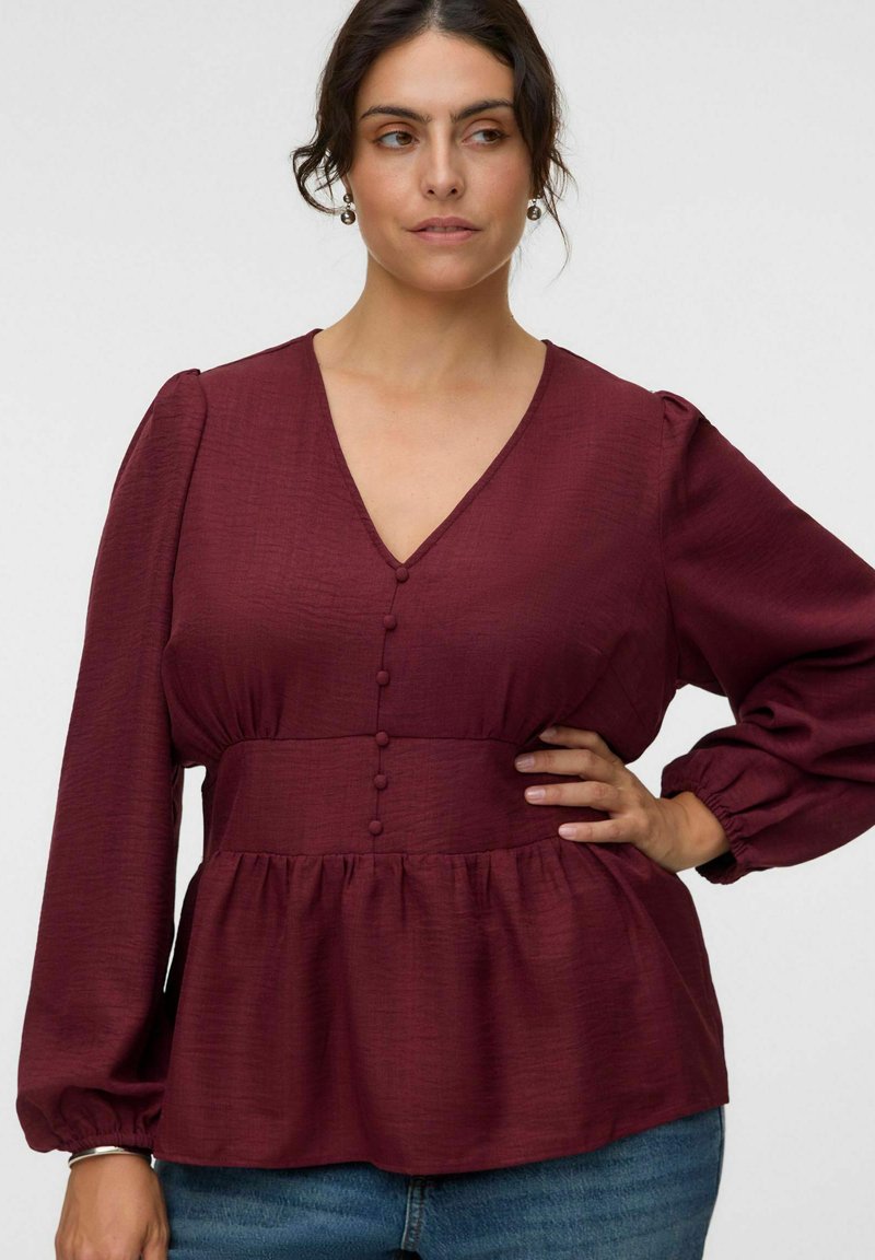 Femme posant la main sur la hanche, portant un chemisier bordeaux à manches longues avec des boutons recouverts de tissu et un jean bleu sur un fond uni.