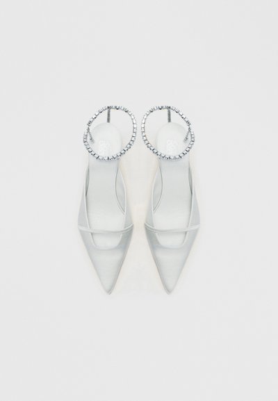 Par de sapatos slingback brancos de bico fino com tiras finas e tiras circulares de tornozelo adornadas com brilhantes, sobre um fundo branco.