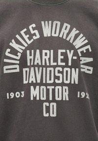 Σκούρο γκρι ύφασμα με λευκό κείμενο που γράφει "Dickies Workwear" τόξο πάνω από "Harley-Davidson Motor Co" και τις χρονιές "1903" και "1920".