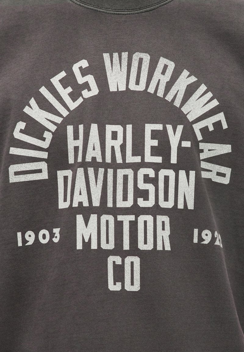 Tessuto grigio scuro con scritta bianca "Dickies Workwear" arcuata sopra "Harley-Davidson Motor Co" e gli anni "1903" e "1920".