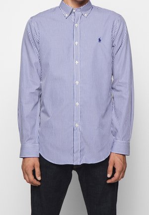 Homme portant une chemise boutonnée à manches longues rayée verticalement bleu et blanc, et un pantalon sombre, debout avec les bras détendus le long du corps.