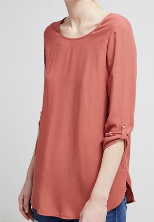 Maglia a maniche lunghe color ruggine con scollatura a barca e polsini arrotolati. Presenta una vestibilità ampia e spacchi laterali per facilitare i movimenti. Texture liscia.