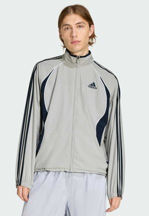 Lichtgrijze trainingsjack met ritssluiting, donkerblauwe accenten, verticale strepen op de mouwen en een opstaande kraag. Voorzien van het Adidas-logo op de borst.