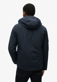 Homme portant une veste à capuche bleu foncé vu de dos sur un fond clair uni.