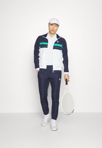 Navy and white athletic tracksuit with teal and black stripes, complemented by a white cap and tennis racket. Smooth fabric, fitted design.