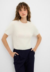Kurzärmliger, kurz geschnittener Pullover in Creme mit strukturierten vertikalen Streifen und rundem Ausschnitt, kombiniert mit dunklen Hosen.
