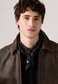 Jeune homme aux cheveux bruns bouclés portant une chemise sombre, une écharpe de cou à motifs navy et une veste en cuir marron, faisant face à l'avant avec une expression neutre.