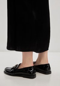 Mocassins vernis noirs avec une finition lisse, un talon bas et des embellissements décoratifs sur le dessus. Présentés sur un fond clair.