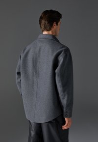 Grå uld skjortejakke med spids krave, knappede manchetter og en glat tekstur. Har en buet kant og detaljer i rygsømmen.