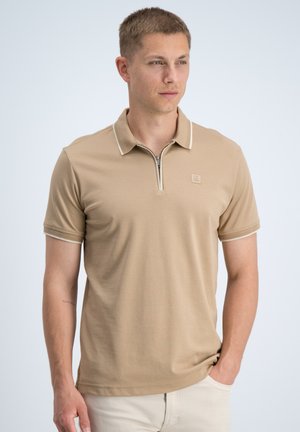 Jonge man draagt een beige polo met korte mouwen en een rits aan de voorkant, kijkt iets naar rechts met één hand in de zak.