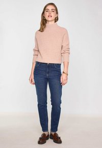 Pull en maille côtelée rose clair avec un col montant, associé à un jean slim bleu foncé et des mocassins marron avec des accents métalliques.