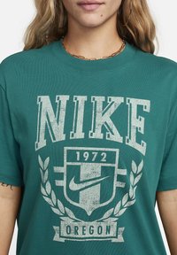 Zielona bawełniana koszulka z wyblakłym białym nadrukiem zawierającym napis "NIKE," "1972," logo Nike oraz "OREGON," otoczone liśćmi laurowymi.