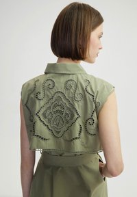 Maglietta verde oliva tagliata con ricami intagliati intricati in un motivo floreale e a spirale. Presenta un colletto e non ha maniche.