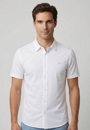 Man draagt een wit overhemd met korte mouwen en kraag, staand tegen een effen lichtgrijze achtergrond.