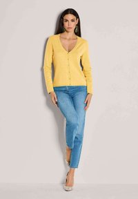 Gelber Cardigan mit V-Ausschnitt, Knopfverschluss und langen Ärmeln. Kombiniert mit hellblauen Jeans und gemusterten High Heels. Weiche Textur.