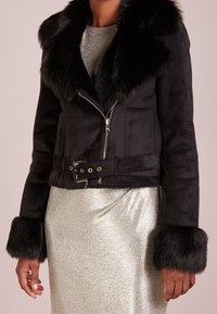 Schwarze Wildlederjacke mit Pelzkragen und -manschetten, tailliert, über einem silbernen metallic Kleid getragen, von Hals bis Hüfte gezeigt.