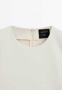 Haut de couleur crème avec un col rond et une fermeture éclair au dos, arborant une étiquette noire avec le texte "Massimo Dutti Studio".