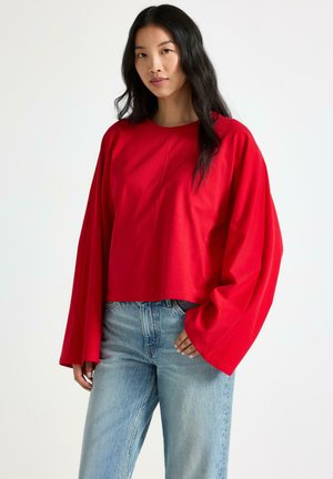 Donna con lunghi capelli neri che indossa una maglia rossa a maniche lunghe e jeans azzurri, in piedi davanti a uno sfondo bianco.