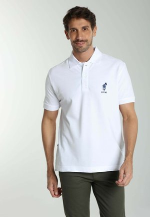 Homme debout et souriant, portant un polo blanc avec un petit logo cheval bleu et un pantalon vert foncé sur un fond uni.