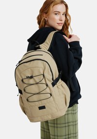 Beige reppu mustilla yksityiskohdilla, etu bungee-köysi, useita vetoketjullisia taskuja, pehmustetut hihnat ja teksturoitu kangaspinta.