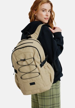 Beige reppu mustilla yksityiskohdilla, etu bungee-köysi, useita vetoketjullisia taskuja, pehmustetut hihnat ja teksturoitu kangaspinta.