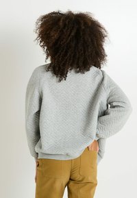 Persona con cabello rizado, vistiendo un sudadera acolchada gris y pantalones mostaza, de pie con una mano en el bolsillo trasero, mirando hacia un lado.